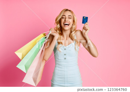 A young woman with long blonde hair is smiling broadly while holding a credit card in one hand and multiple shopping bags in the other. She is wearing a light blue dress against a pink background. A young woman with long blonde hair is smiling broadly while holding a credit card in one hand and multiple shopping bags in the other. She is wearing a light blue dress against a pink background. 123364760