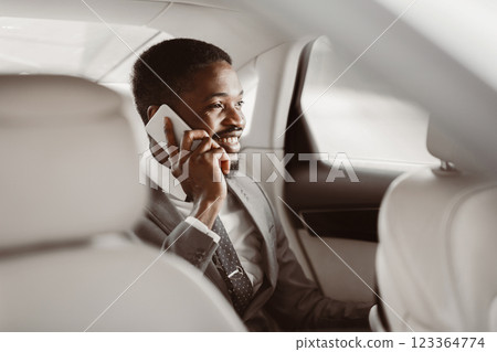 Black businessman in a suit sits in the back seat of a car and talks on his cell phone. He is smiling and appears to be enjoying the conversation. The cars interior is clean and modern. 123364774