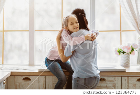 A young girl wearing jeans and a pink shirt hugs her mother from behind while standing near a window in a kitchen. They both appear to be smiling. 123364775