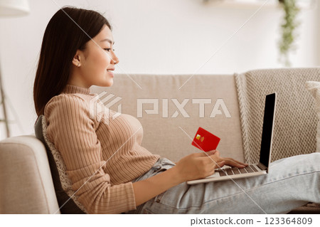 A young woman sits on a couch, looking intently at a laptop as she holds a credit card, likely making an online purchase. 123364809