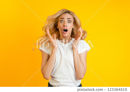 Young woman with long blonde hair wearing a white t-shirt looking shocked in front of a yellow background. Her mouth is open, her eyes are wide, and her hands are raised in a gesture of surprise. Young woman with long blonde hair wearing a white t-shirt looking shocked in front of a yellow background. Her mouth is open, her eyes are wide, and her hands are raised in a gesture of surprise. 123364819