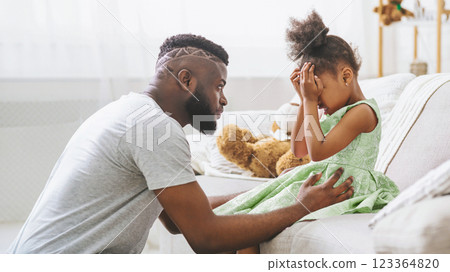 Black father is kneeling on a couch beside his young daughter, who is crying and covering her face with her hands. He is reaching out to her with a comforting hand on her stomach. 123364820