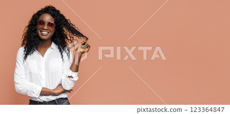 A smiling Black woman with long curly hair is wearing sunglasses and a white button-down shirt. She is standing with her arms crossed in front of a peach-colored background A smiling Black woman with long curly hair is wearing sunglasses and a white button-down shirt. She is standing with her arms crossed in front of a peach-colored background 123364847