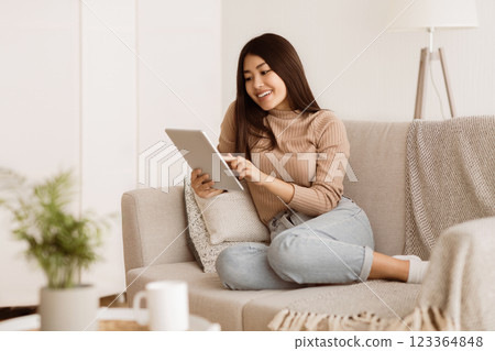 A woman sits on a couch at home, using a tablet. She is smiling and appears to be relaxed. 123364848