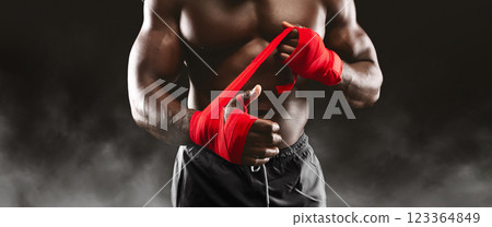 A muscular boxer prepares for a fight by wrapping his hands with red tape. A muscular boxer prepares for a fight by wrapping his hands with red tape. 123364849
