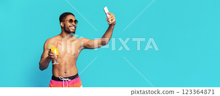 A young black man in swimwear poses for a selfie while holding a cocktail. He is wearing sunglasses and smiling. The background is a solid light blue, copy space 123364871