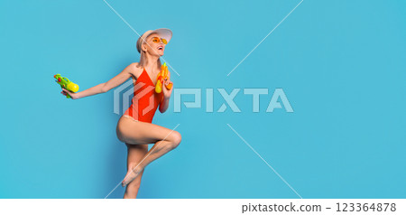 A woman in a red swimsuit and a white sun hat stands with one leg raised and poses for the camera while holding two water guns in front of a bright blue background, copy space A woman in a red swimsuit and a white sun hat stands with one leg raised and poses for the camera while holding two water guns in front of a bright blue background, copy space 123364878