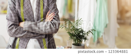 A woman stands in a design studio, wearing a plaid blazer with a yellow measuring tape hanging around her neck. 123364881