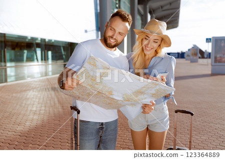 Smiling couple checking location using smart phone and paper map standing on the street with suitcases 123364889