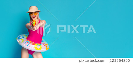 A young woman is wearing a pink swimsuit and a straw hat, standing with a bright yellow water gun in her hand. She is holding an inflatable ring in front of her and is smiling broadly. 123364890