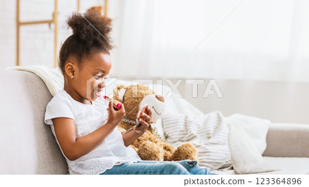 African American young girl with an afro hairstyle is sitting on a couch, applying lipstick while looking in a small mirror. She is wearing a white t-shirt and blue jeans, copy space 123364896