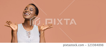 African American young woman with short, dark hair is smiling brightly while standing against a peach-colored background. She has her arms extended outwards with her palms open and facing the viewer 123364898