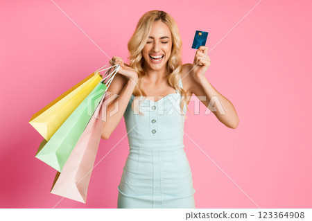 A young woman with blonde hair is holding multiple shopping bags and a credit card while smiling and looking happy against a pink background. 123364908
