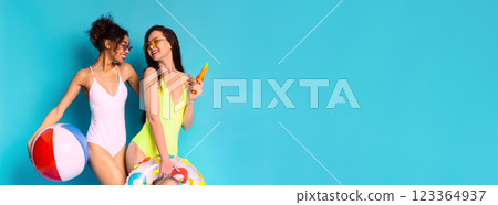 Two young women are posing in front of a blue background. Both women are wearing swimsuits and sunglasses and are smiling at the camera, copy space 123364937