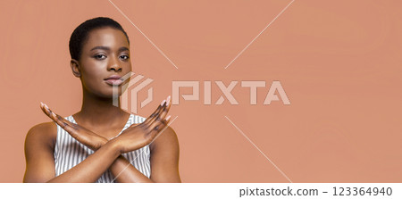 African American young woman with short black hair is standing in front of peach background. She is wearing a white and black striped tank top, crossing hands in front of chest, showing stop gesture African American young woman with short black hair is standing in front of peach background. She is wearing a white and black striped tank top, crossing hands in front of chest, showing stop gesture 123364940