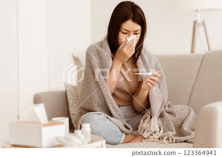 A woman wrapped in a blanket sits on a couch and checks her temperature with a thermometer while holding a tissue to her nose. 123364941