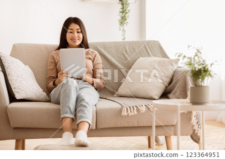 A woman sits on a couch in a living room, relaxed and browsing on a tablet. 123364951
