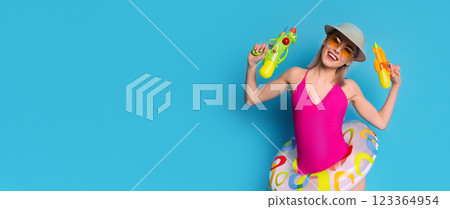 A young woman in a pink swimsuit, a sun hat, and sunglasses holds two water guns. She smiles at the camera with a playful expression. Her joyful demeanor suggests fun and excitement 123364954
