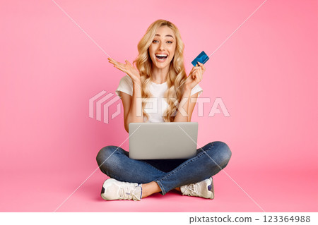 A young blonde woman sits cross-legged on a pink background, smiling excitedly as she holds a credit card and uses a laptop. 123364988