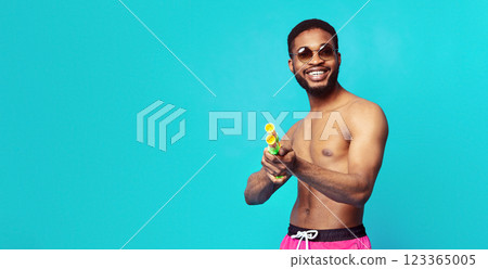 A smiling African American man in swim trunks and sunglasses is holding a water gun in front of a blue background, copy space 123365005