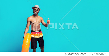 African American man in swim trunks, sunglasses, and a straw hat smiles and gives a thumbs up while holding an inflatable ring in front of a blue background. 123365026