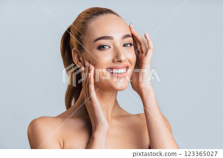 A young woman with long brown hair tied back in a ponytail smiles brightly at the camera. She is touching her face with both hands, and her shoulders and chest are visible. 123365027