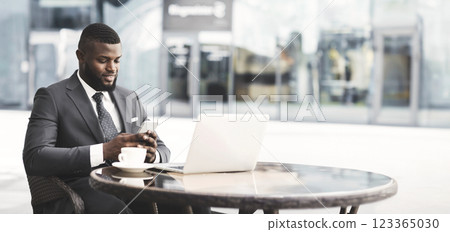 Business Lifestyle. Smiling Businessman Using Cellphone Texting Working On Laptop Sitting In Outdoor Cafe In Urban Area Business Lifestyle. Smiling Businessman Using Cellphone Texting Working On Laptop Sitting In Outdoor Cafe In Urban Area 123365030