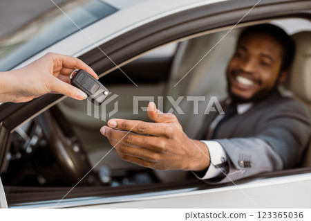 African American man is sitting in the drivers seat of a car with a person handing him the keys. He is smiling and reaching out to take them. African American man is sitting in the drivers seat of a car with a person handing him the keys. He is smiling and reaching out to take them. 123365036