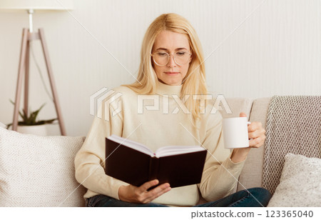 A woman wearing glasses sits on a couch, reading a book and holding a cup of coffee. 123365040