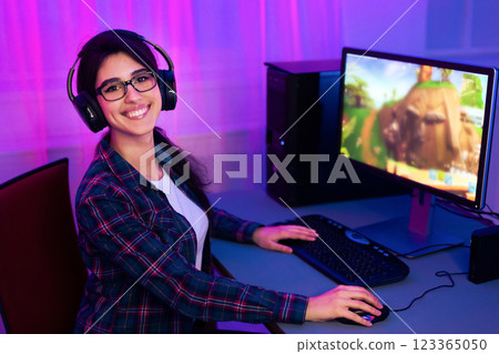 A young woman with long dark hair is smiling as she plays a computer game. She is wearing a plaid shirt and headphones. The computer monitor in the background shows an animated video game. A young woman with long dark hair is smiling as she plays a computer game. She is wearing a plaid shirt and headphones. The computer monitor in the background shows an animated video game. 123365050