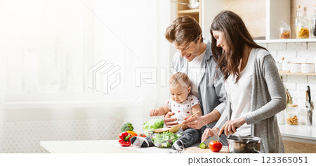 A family of three is cooking together in their kitchen. The father is holding their baby, who is sitting on the counter and reaching for a wooden spoon, copy space A family of three is cooking together in their kitchen. The father is holding their baby, who is sitting on the counter and reaching for a wooden spoon, copy space 123365051
