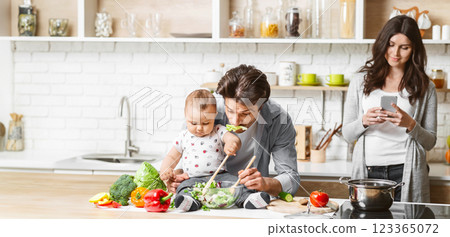 A father feeds his baby a salad while sitting at the kitchen counter. The baby is happily eating the salad, while the father smiles at the baby. The mother stands behind them, copy space 123365072