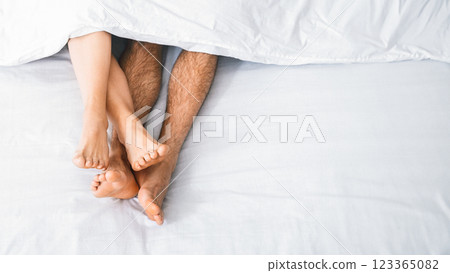 A close-up image of a couples bare feet intertwined under a white sheet, visible from the top of the bed. The focus is on the feet, with the sheet providing a contrasting background. 123365082