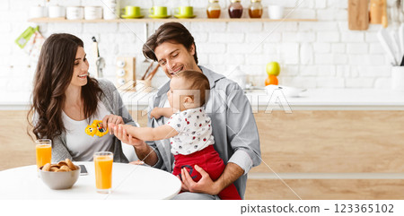 A family of three, a mother, a father, and a baby boy, is enjoying a breakfast meal together in their bright and modern kitchen. The father is holding the baby in his arms, copy space 123365102