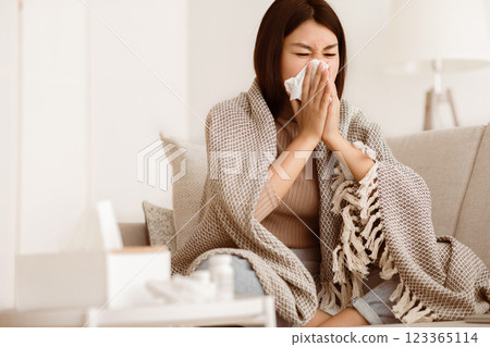 A woman sits on a couch wrapped in a blanket, blowing her nose with a tissue. A woman sits on a couch wrapped in a blanket, blowing her nose with a tissue. 123365114