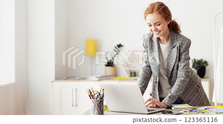 A woman in a blazer smiles as she works on a laptop in a modern office. 123365116