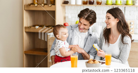 A happy family is enjoying breakfast together in a kitchen. The father is holding their baby, who is smiling and looking at the father. The mother is smiling and looking at the baby A happy family is enjoying breakfast together in a kitchen. The father is holding their baby, who is smiling and looking at the father. The mother is smiling and looking at the baby 123365118