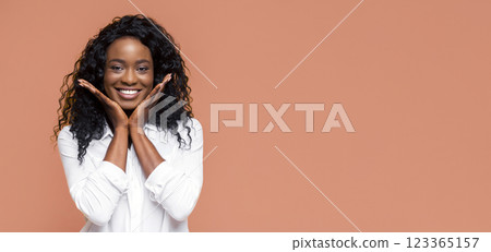 A smiling black woman with curly hair poses with her hands on her cheeks in front of a peach-colored background. She is wearing a white button-down shirt. 123365157