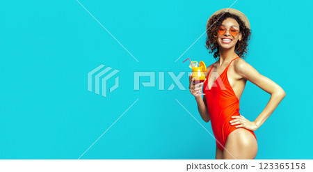 A smiling black woman in a red swimsuit and a straw hat is holding a tropical cocktail with a straw. She is wearing orange sunglasses and standing against a bright blue background. 123365158
