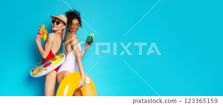 Two women in swimsuits are posing with pool floats and a water gun on a blue background. They are smiling and seem to be having a good time, copy space Two women in swimsuits are posing with pool floats and a water gun on a blue background. They are smiling and seem to be having a good time, copy space 123365159