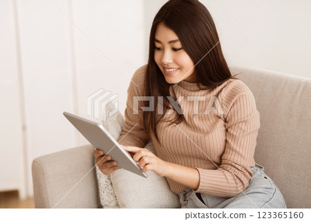 A young woman smiles as she scrolls through her tablet while sitting on a couch. 123365160