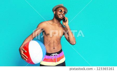 A young, smiling black man is wearing sunglasses and a hat, standing against a bright blue background. He is holding a beach ball in his left hand and talking on a phone with his right hand A young, smiling black man is wearing sunglasses and a hat, standing against a bright blue background. He is holding a beach ball in his left hand and talking on a phone with his right hand 123365161
