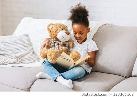 A young black girl with curly black hair sits on a light gray couch, holding a book open in her lap while wearing a white shirt and blue jeans. A young black girl with curly black hair sits on a light gray couch, holding a book open in her lap while wearing a white shirt and blue jeans. 123365162