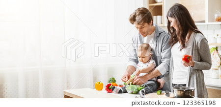 A family of three is preparing food together in a kitchen. The father is holding their baby while they all help prepare vegetables. The mother is standing next to them, copy space A family of three is preparing food together in a kitchen. The father is holding their baby while they all help prepare vegetables. The mother is standing next to them, copy space 123365198