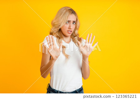 A woman with blonde hair stands in front of a yellow background, showing both hands in a stop gesture. She has a concerned expression on her face, trying to deter someone from approaching. A woman with blonde hair stands in front of a yellow background, showing both hands in a stop gesture. She has a concerned expression on her face, trying to deter someone from approaching. 123365221