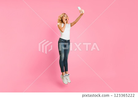 A young blonde woman wearing a white shirt and blue jeans is jumping in mid-air against a pink background. She is holding a smartphone in her right hand and taking a selfie 123365222