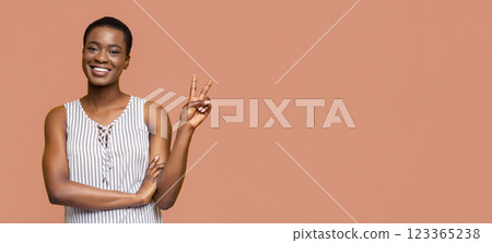 African American young woman with short hair is smiling and giving a peace sign with her right hand. She is wearing a white and black striped sleeveless top and has her left arm crossed over her torso African American young woman with short hair is smiling and giving a peace sign with her right hand. She is wearing a white and black striped sleeveless top and has her left arm crossed over her torso 123365238