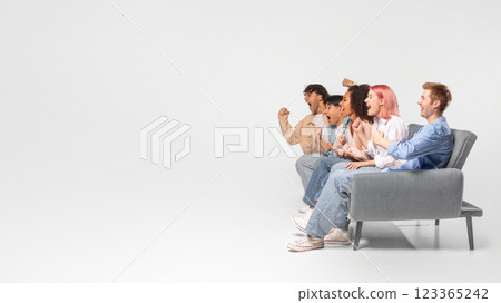 A group of five friends sit on a gray couch, excitedly watching a television screen that is off-camera. The friends are all dressed casually and have a variety of hairstyles, copy space A group of five friends sit on a gray couch, excitedly watching a television screen that is off-camera. The friends are all dressed casually and have a variety of hairstyles, copy space 123365242