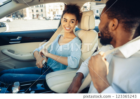 A young black woman and man in the front seats of a car are buckling up their seatbelts. They are smiling and appear to be enjoying their time together. The car is modern and has a sleek interior. 123365243