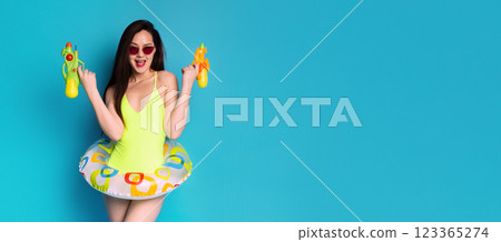 A happy woman in a bright yellow swimsuit is smiling at the camera. She is wearing sunglasses, holding a water gun in each hand, and has a colorful inflatable ring around her waist, copy space A happy woman in a bright yellow swimsuit is smiling at the camera. She is wearing sunglasses, holding a water gun in each hand, and has a colorful inflatable ring around her waist, copy space 123365274